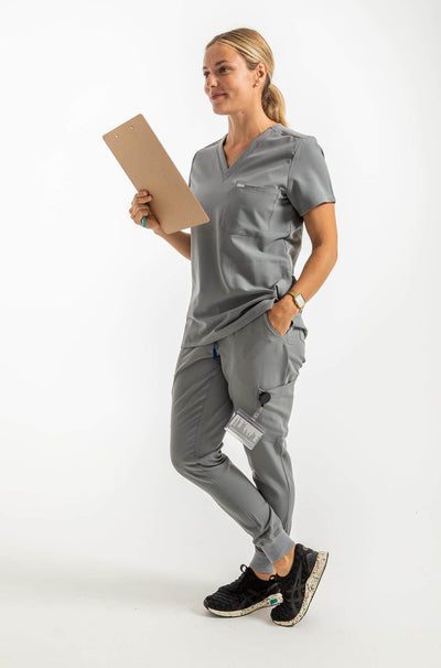 VENA ladies jogger style scrub shirt  lady holding checklist and 1 hand in pant side pocket#colour_grey